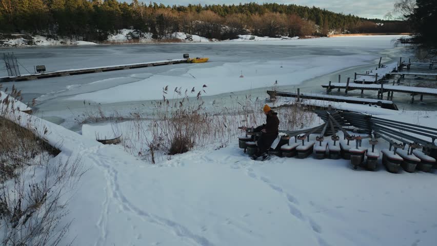 A drone shot of a man with yellow hat leaning on a fence and drinking something in snow-covered area with forest in the background