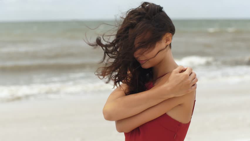 A pensive walking woman in a red dress embraces herself on a windy beach, lost in thought. Perfect for concepts of loneliness, contemplation, and introspection in travel, fashion, and lifestyle