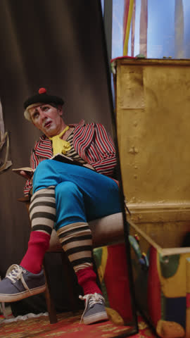 Full vertical shot of thoughtful male clown reflecting in mirror sitting on chair in circus backstage area, wondering and writing in notebook