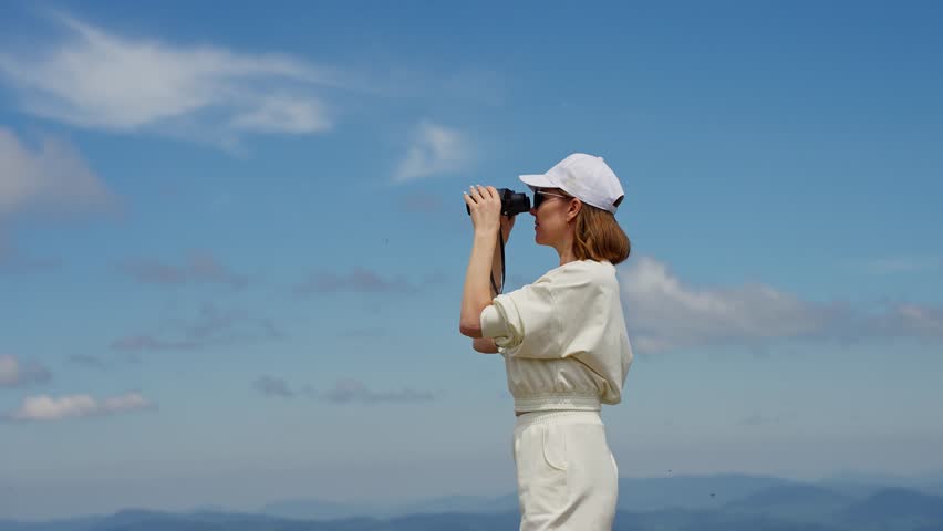 A woman in white clothing stands atop a mountain and uses binoculars to survey the expansive landscape. She looks out at the horizon with a sense of freedom and wonder.
