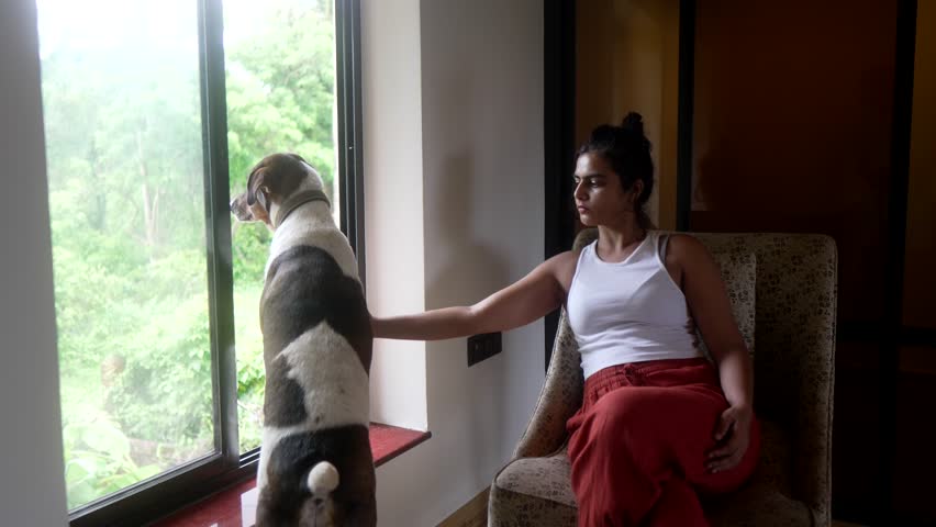 Woman in white tank top pets dog while sitting by window, looking out at lush green scenery