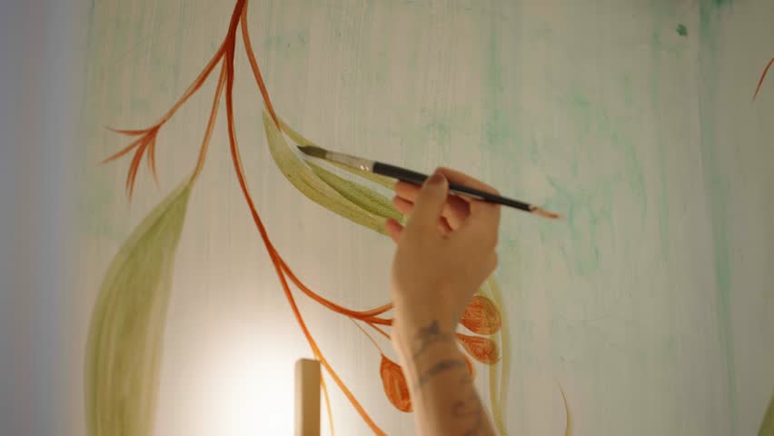 An artist applies colorful acrylic paint to a white wall with a brush. A young woman artist works in her workshop. Nature painting, contemporary art.
