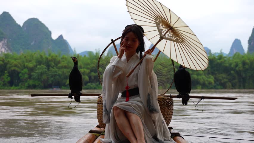 Hanfu girl seated on a bamboo raft with two cormorant birds on sides by Li River