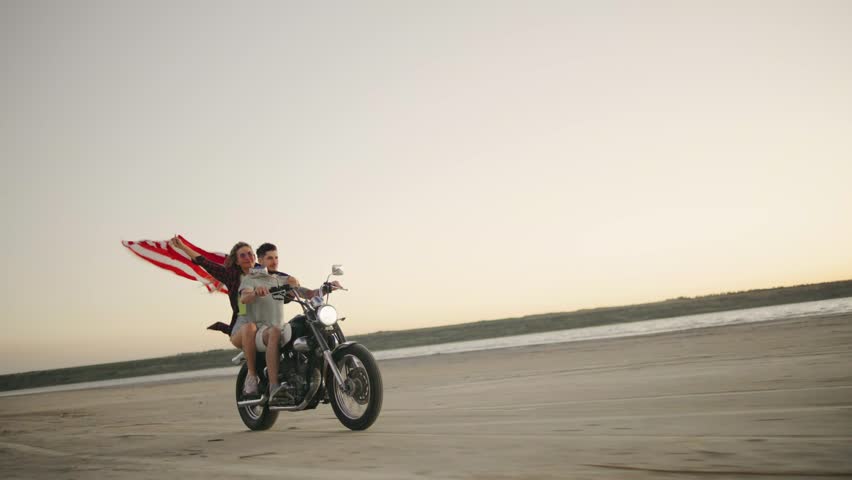 A couple in love with an American flag rides a motorcycle on the beach during sunset, enjoying a moment of freedom - Powered by Shutterstock - Get 15% off with code: PIKWIZARD15