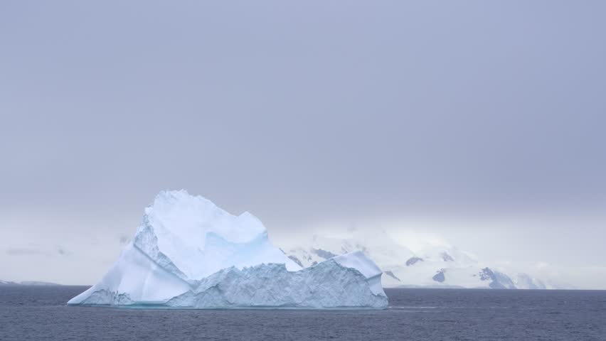Large Iceberg in Antarctica. Pass by Floating Ice Blue Ocean White Snow Mountains in Background