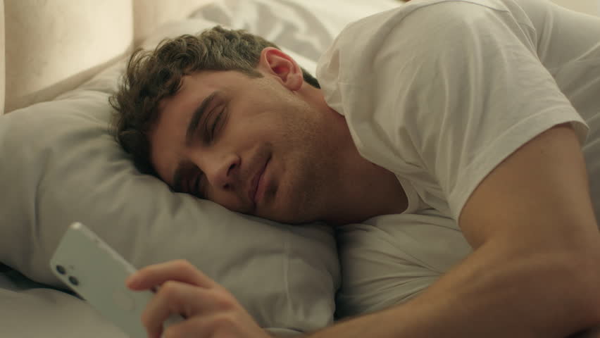 Caucasian man wake up looking mobile phone smartphone telephone gadget sleepy continue sleep lying on bed male weekend check time lazy daydream slumber nap resting technology device guy home bedroom - Powered by Shutterstock - Get 15% off with code: PIKWIZARD15