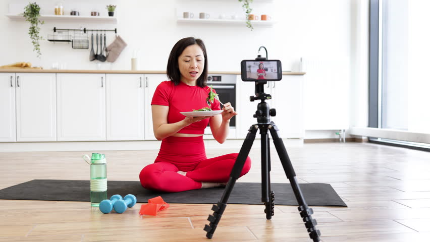 Woman in red activewear sitting on yoga mat eating salad while filming fitness video in modern kitchen. Healthy lifestyle, nutrition, and social media influencer concept.