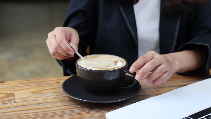 A young, attractive Asian businesswoman sits in a cafe, holding a mug of hot coffee, smiling. She