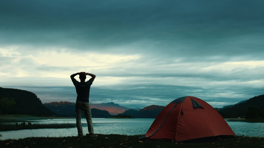 Hiker standing by the lake and watching mountain on the horizon with camping tent on the foreground