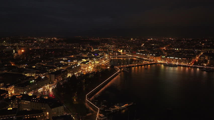 Aerial view of Geneva's lit skyline and waterfront at night, capturing Jardins des Anglais and traffic over Pont du Mont Blanc