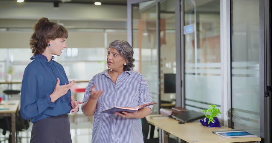 Indian serious old business women standing hold book make new project discussion indoor office room. Beautiful young adult corporate expert girl staff team do work talk day task share ideas job place