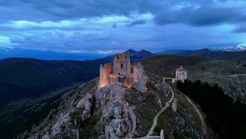 A drone of illuminated Rocca Calascio castle in Abruzzo, Italy with the Church of the Madonna della Pieta on top of mountain at cloudy evening