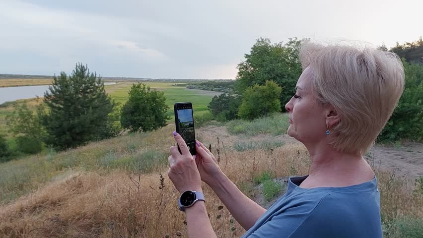 A beautiful woman takes photos and videos of nature, landscape and flowers on her mobile phone on a summer day