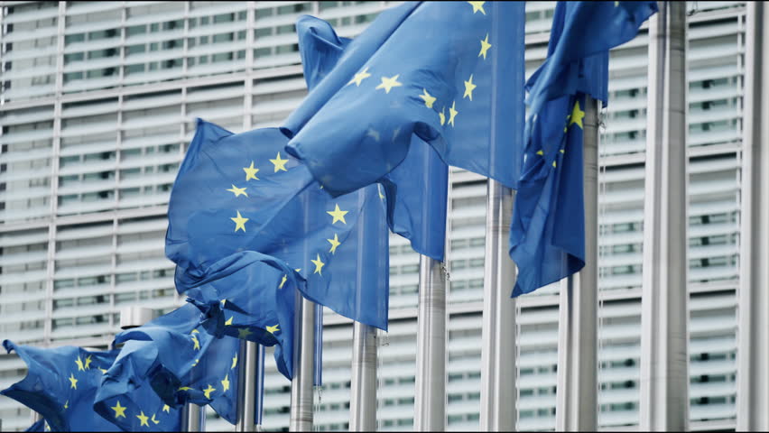 In Brussels, the political heart of the European Union, several EU flags flutter in harmony beside a contemporary glass building, embodying unity and progressiveness in a symbolism rich scene