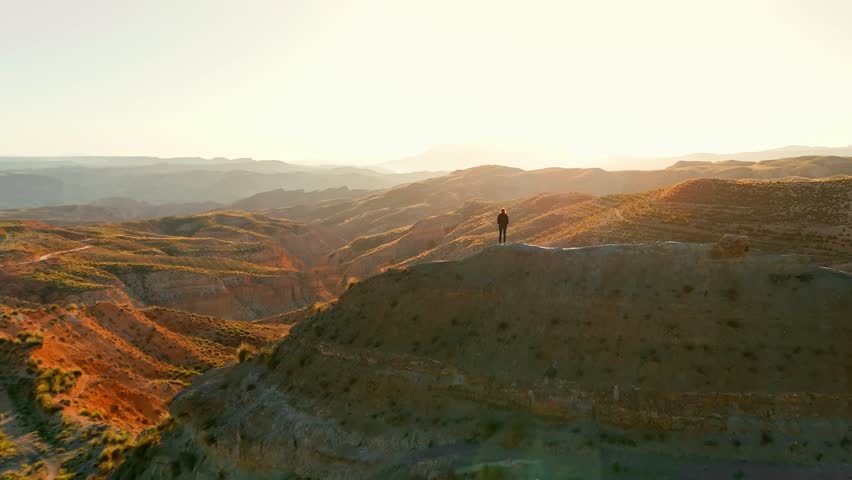 Young woman standing on top of mountain summit at sunset. Watching amazing sunset over the mountainous desert landscape of Gorafe desert in Andalusia, Spain. Aerial drone footage.