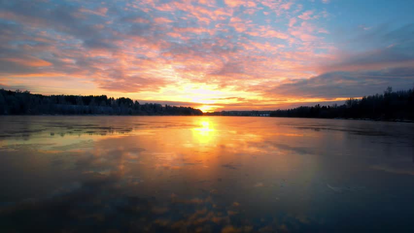 A drone shot of a frozen lake in a forest with a scenic cloudy dusk sky in the background