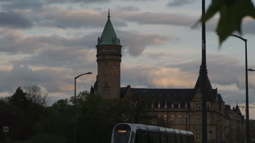 Modern tram arrives at the station against the medieval brick buildings with spiers in Luxembourg during sunset. Free public transportation in a beautiful city Luxembourg