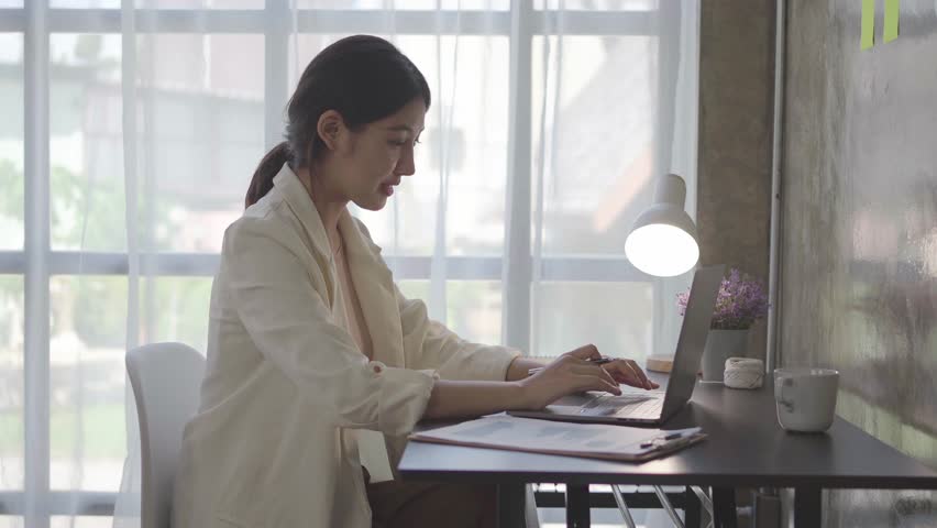Focused young businesswoman works on her laptop in a modern office, typing and taking notes. She appears productive and professional in her work environment