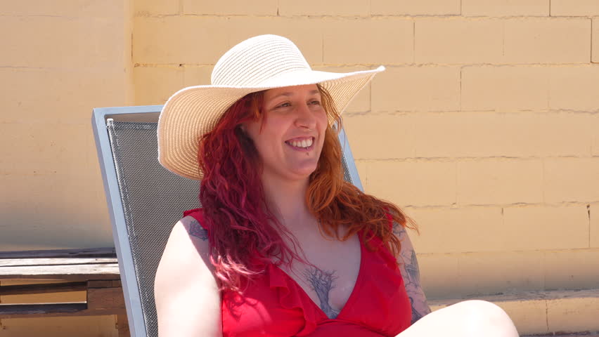 A woman in a red bikini is sitting on a chair with a straw hat on her head. She is smiling and she is enjoying her time