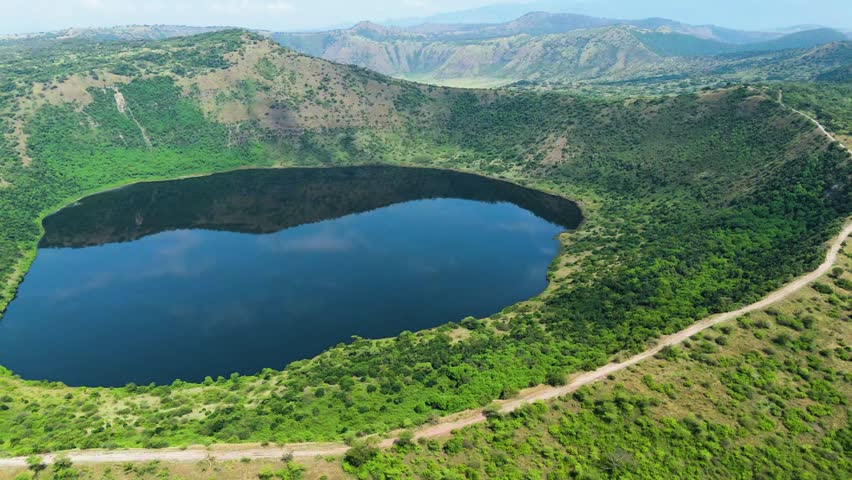 Breathtaking aerial drone footage of a pristine crater lake surrounded by lush green hills and winding paths in Uganda. Captures the serene beauty and unique landscape of the region.
