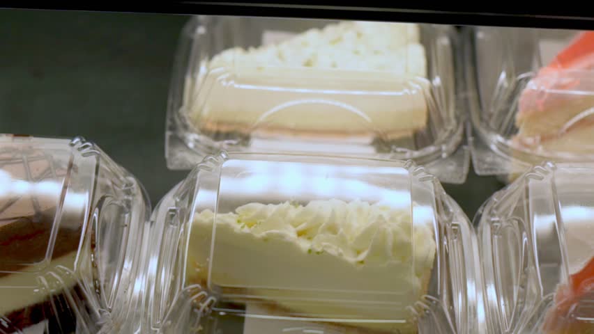 Close-up of various dessert slices, including cheesecake and chocolate cake, packaged in clear plastic containers, displayed in a store.