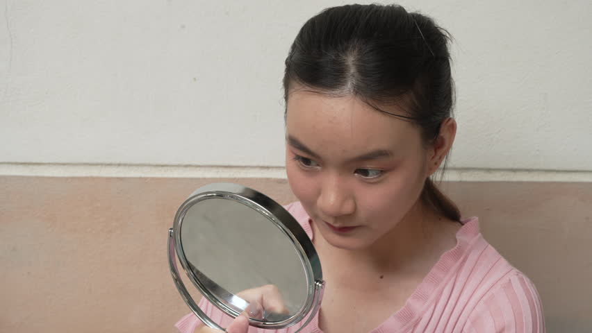 A young Asian woman in a pink dress sits on the front house stairs, holding a mirror and looking at her own face, enjoying a moment of self-reflection and beauty.