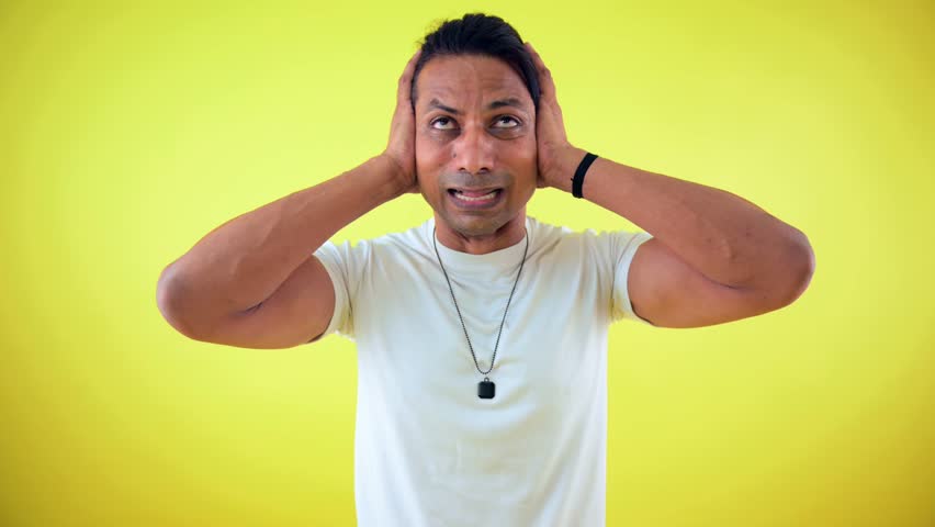 Fearful man crying and covering ears, Young person shed tears in fear, Men apprehensive expression, is scared badly, Afraid male cry while upset, White t-shirt, on yellow background, Indian, bowl, 30s