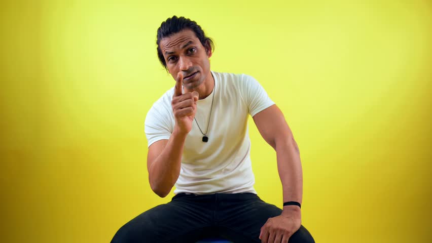 Young man sits with aggressive expression, Person scolding someone while sitting, is threatening on camera, Looking very angry and arrogant, A men in white t-shirt, against yellow background, Indian
