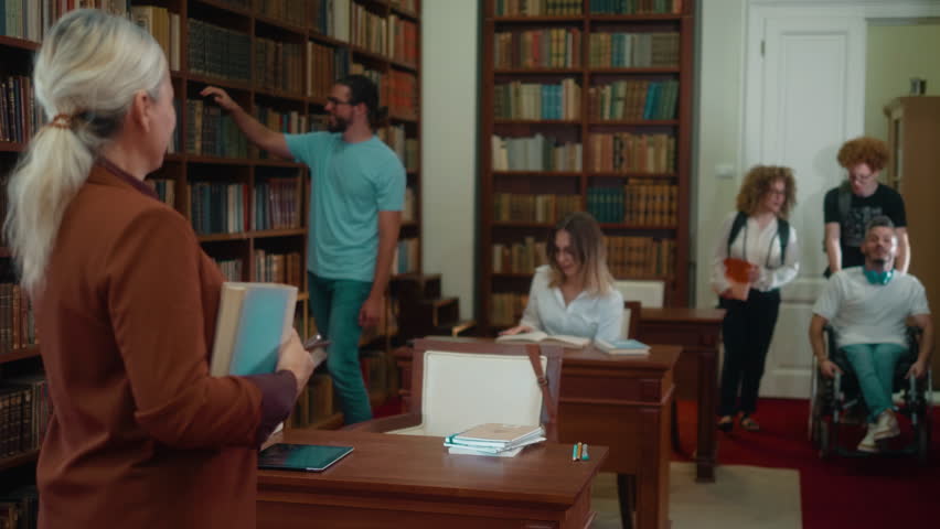 A group of students entering a university classroom where a lecturer is waiting for them. Female student enters the classroom for class pushing her colleague in a wheelchair