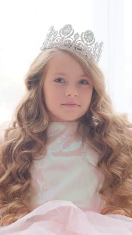 Portrait of a cute little princess with a crown on her head. The girl winks and smiles at the camera. Vertical video.