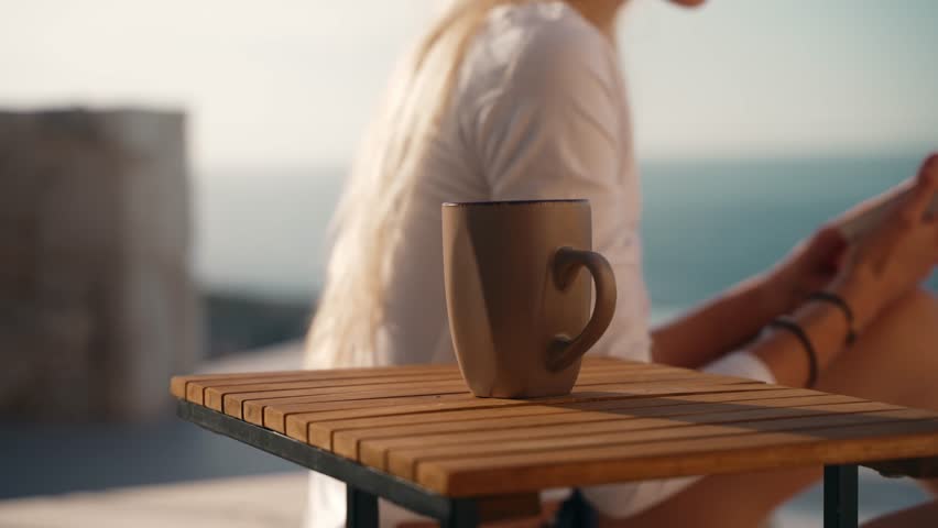 Young caucasian woman taking a cup of coffee from a table while reading a book outside during summer