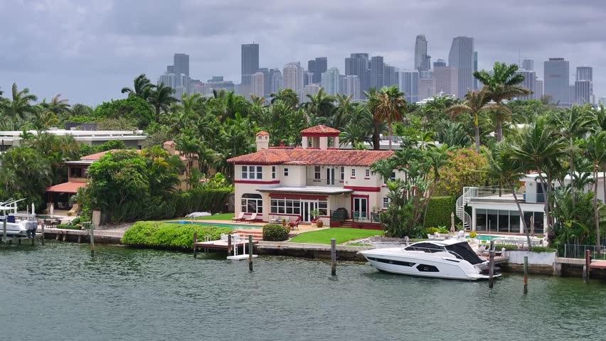 Expensive waterfront mansions on Venetian Islands with Miami skyline in background. Aerial parallax shot revealing sprawling cityscape of wealthy American urban metropolis.