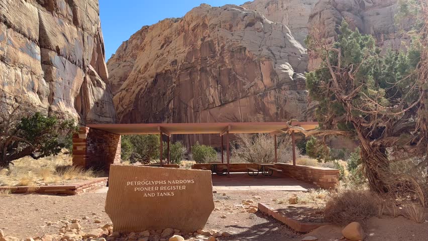 Capitol Reef National Park trailhead. A picnic area sits before a sandstone canyon wall and a trail sign. The sign points to the Petroglyphs Narrows, Pioneer Register and Tanks trails - Utah, USA
