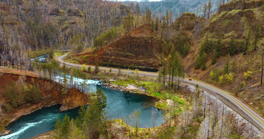 Flying closer to the highway at the foot of the mountains. Red car rides by the road along the rock. Oregon nature.