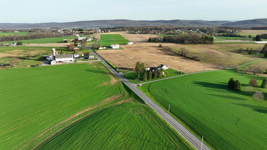 Drone flyover farm house with silo surrounded by cultivation fields in american countryside. Sunny day in the evening. Car on rural road in suburb.