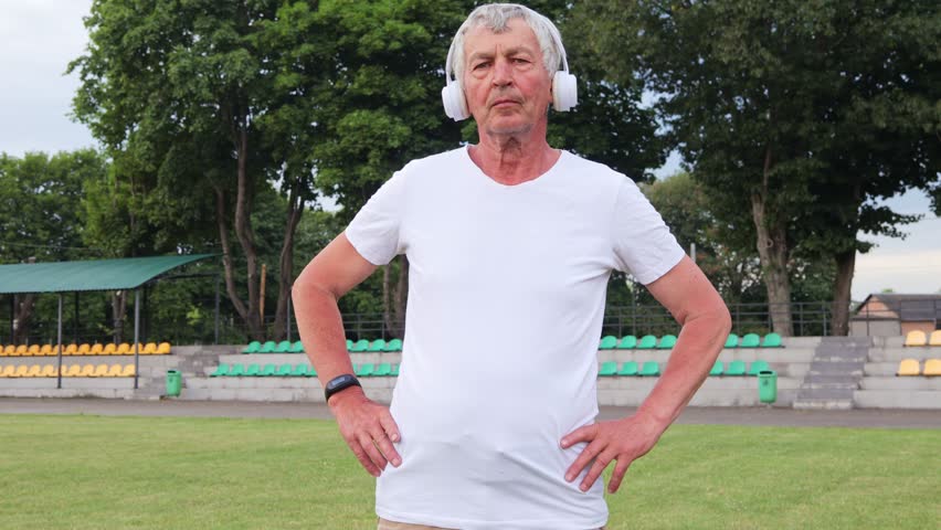 Retired male Caucasian pensioner wearing white T-shirt and headphones enjoying healthy lifestyle by engaging in stretching and warming up before his workouts