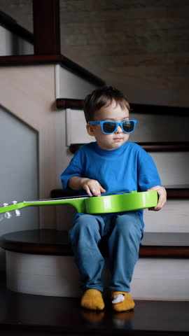 Little lovely kid wearing glasses sits on the stairs. Baby boy holding a small guitar on his knees. Vertical video.