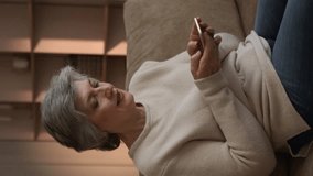 Vertical Screen: senior woman relaxing on a couch at home while using her smartphone. She appears to be scrolling through something on her phone, perhaps looking at photos, watching videos, or reading