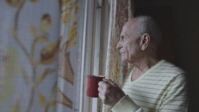 Elder man holding red mug looks out of window with floral curtains. He wears yellow striped sweater and appears thoughtful. - Powered by Shutterstock - Get 15% off with code: PIKWIZARD15