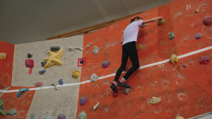 A little girl of 10 years old, a professional climber, trains on a climbing wall indoors. Climbs at high altitude and sits on the splits to reach the ledge on the wall. High quality 4k footage