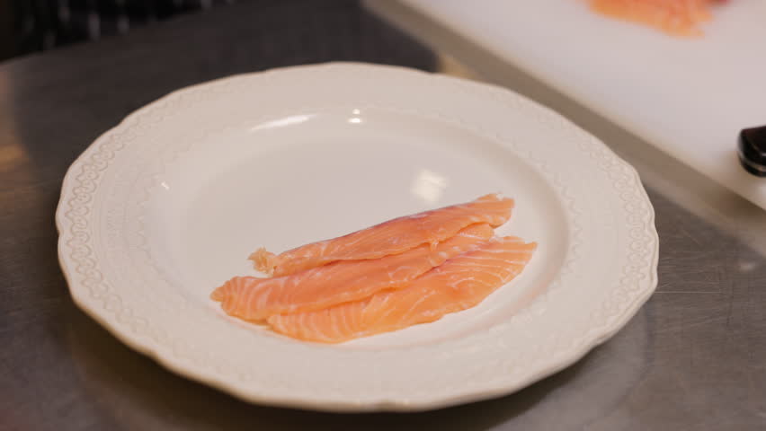 Hands of competent culinary expert placing slices of smoked salmon onto white plate with embossed edge. Presentation of fresh seafood on kitchen environment.