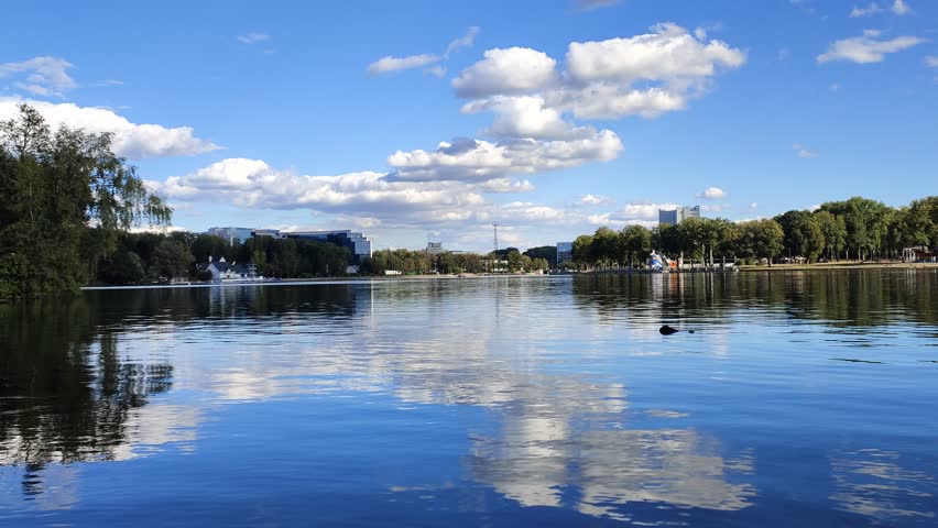 Birch trees and other trees grow on the shores of the lake. On the far shore - the embankment and city buildings. The water is rippling Blue sky with clouds reflected in the water Sunny autumn weather