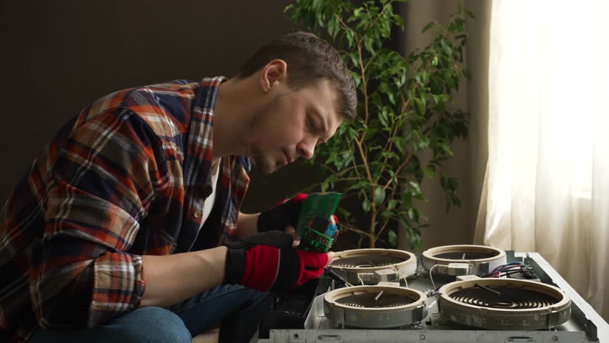 Man technician in gloves examines industrial fan parts against plant in workshop. Young guy checks details of device discovering smell of burning. Experienced male worker prepares for repair procedure