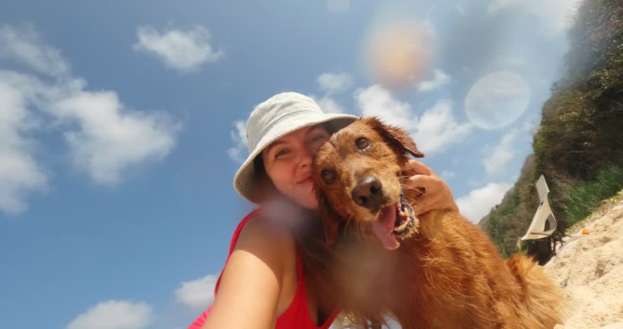 A young woman in a red swimsuit on the beach takes a selfie with her golden retriever dog. She hugs the dog and kisses its face. Hiking with tents and a dog during summer vacation.