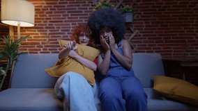 Two multiracial young women sitting on a couch, looking scared while watching a horror movie. Two Young Women Watching a Scary Movie on Couch - Powered by Shutterstock - Get 15% off with code: PIKWIZARD15