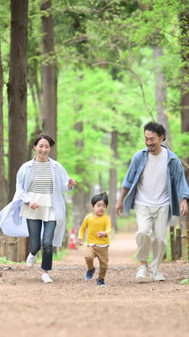 A happy Japanese family running amicably through fresh greenery and beautiful green parks. Three smiling people. Images of insurance, leisure and outings. Vertical video.