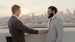 Two businessmen from different backgrounds are having a discussion about finances and seal their agreement with a handshake on a rooftop at sunset, with a city skyline in the background - Powered by Shutterstock - Get 15% off with code: PIKWIZARD15