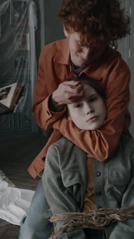 Vertical shot of young guy drawing cross with smouldering palo santo stick on forehead of devil possessed girl tied up to chair with rope during exorcism rites in derelict mansion