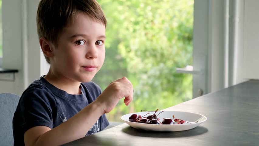 6 year cute little preschool boy eating large ripe cherries. Happy child holding in hand fresh cherry. Healthy organic berries on plate. Summertime season. In the kitchen at the table. Delicious food.