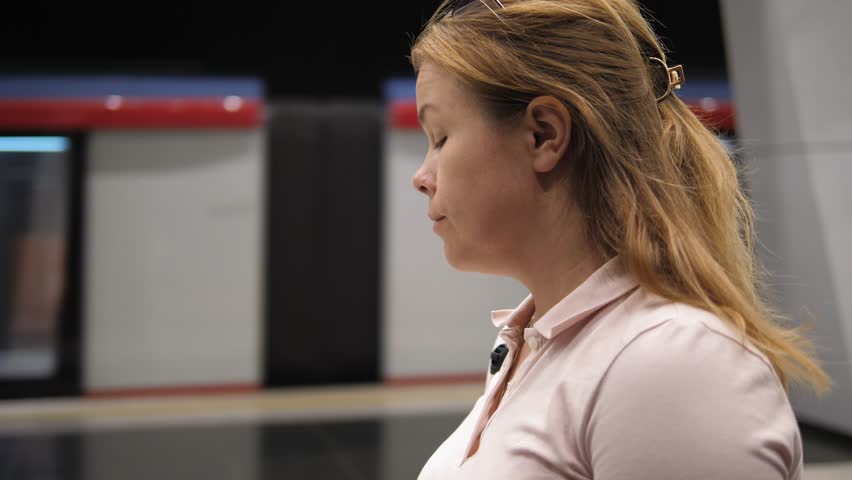 Side view of a young woman with a backpack at the metro station, the train is moving along the rails. A woman with a phone in the subway is waiting for a train and looking around.